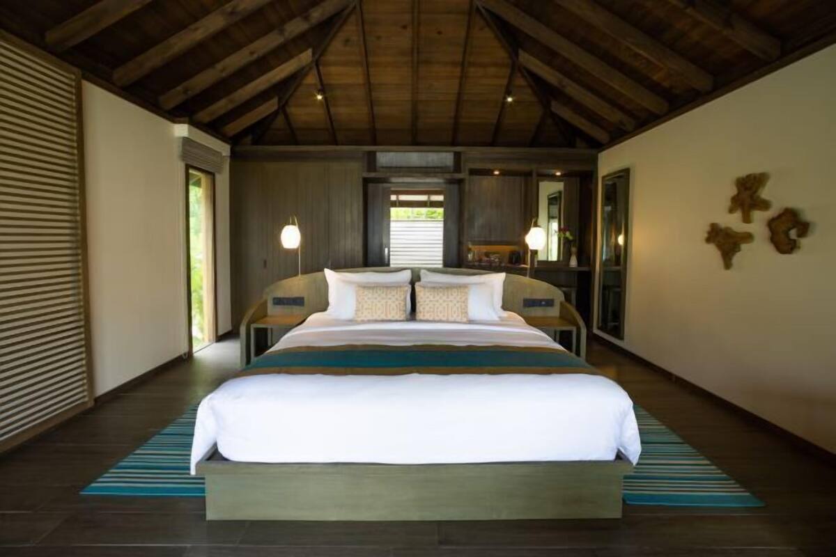 A bedroom with a large bed, wooden ceiling beams, a window, and a nightstand with a lamp.