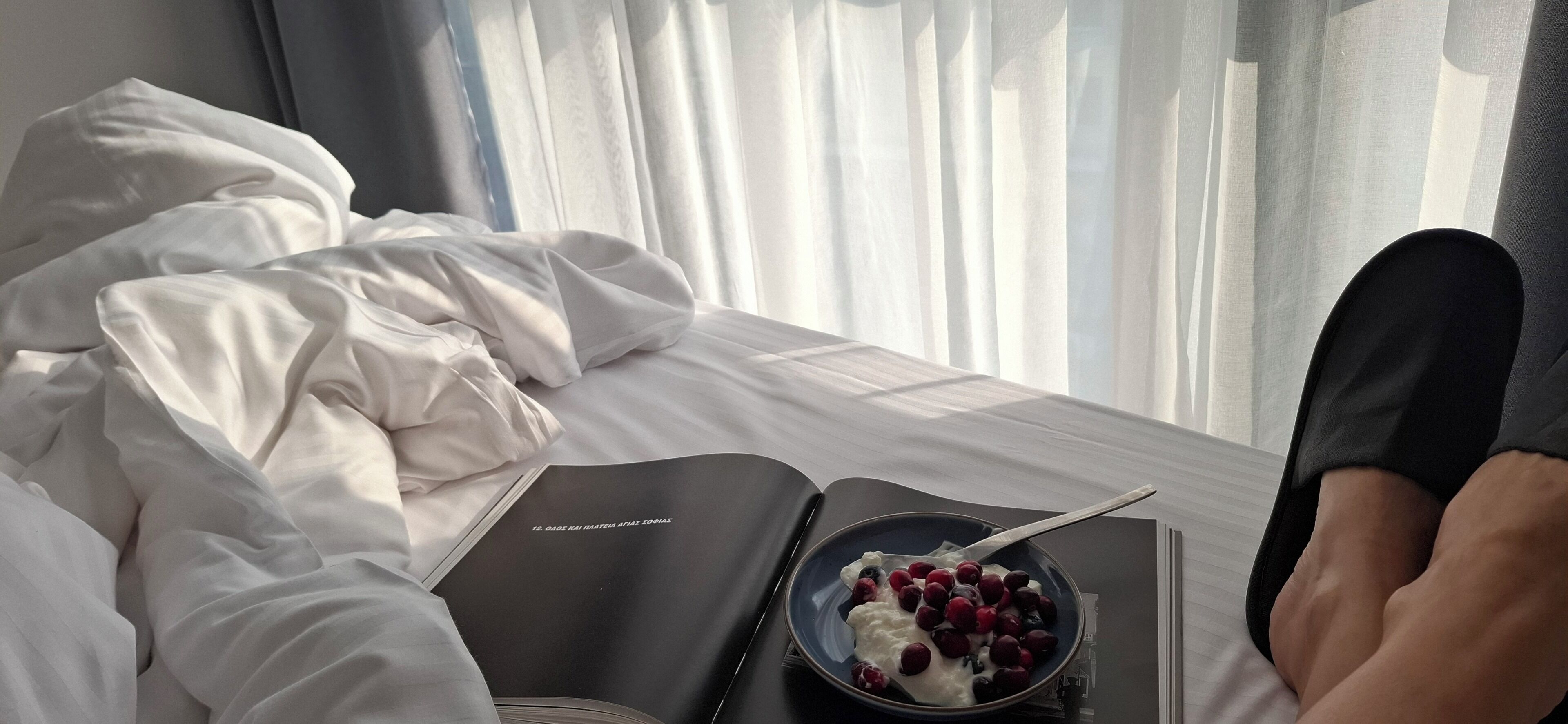 A bed with white linens, a book, and a bowl of yogurt with berries on a tray.