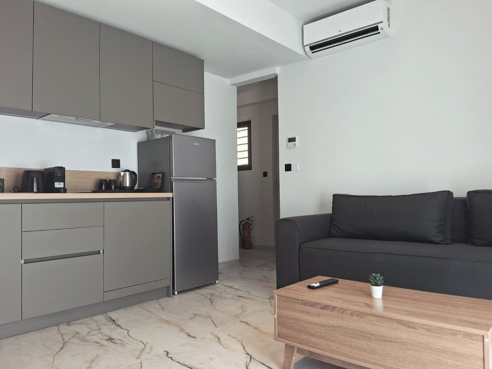 Cuina moderna amb armaris grisos, un refrigerador d'acer inoxidable i una cafetera. Hi ha un sofà gris fosc i una taula de centre de fusta amb un comandament a distància i una petita planta.