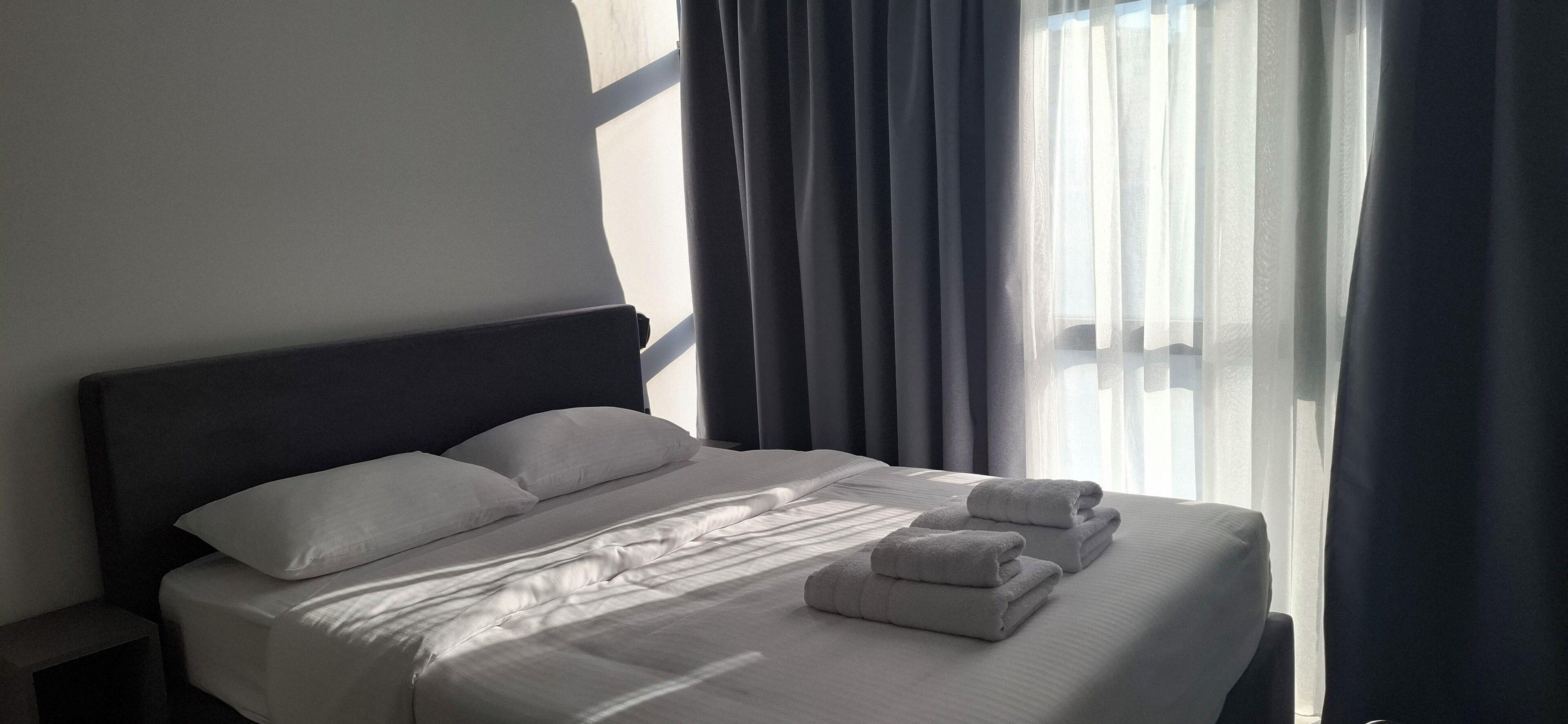 Un lit bien fait, avec du linge de lit et des serviettes blanches, une tête de lit sombre et une vue sur l’extérieur à travers des rideaux légers.
