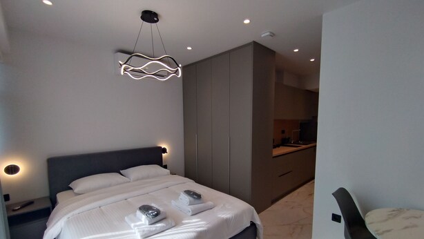 Une chambre à coucher moderne avec un grand lit, des tables de chevet, un luminaire suspendu et une vue sur la cuisine.