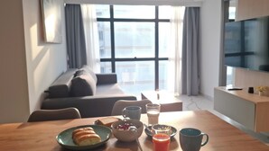 A modern living room with a sofa, a dining table with breakfast items, a TV, and a large window with curtains. - Vardaris Apartments (Thessaloniki)