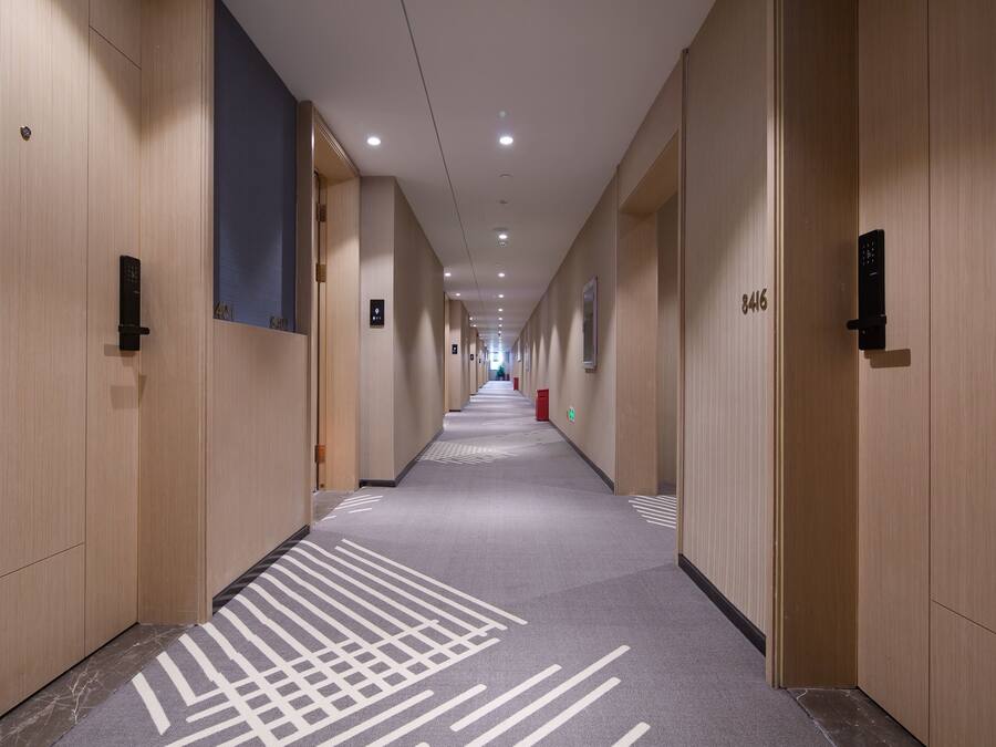 Un largo y estrecho pasillo de hotel con paredes de madera, puertas numeradas y una alfombra estampada.