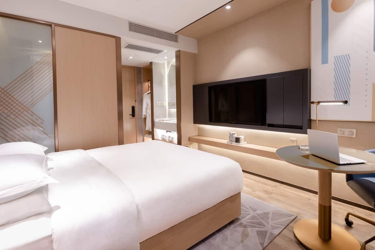 Ein modernes Hotelzimmer mit einem großen Bett, einem Schreibtisch mit einem Laptop, einem Fernseher und einem durch eine Glastür einsehbaren Badezimmer.