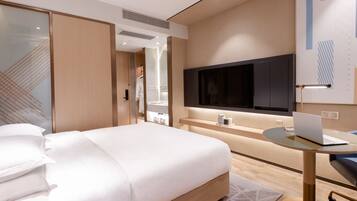 A modern hotel room with a large bed, a desk with a laptop, a TV, and a bathroom visible through a glass door.