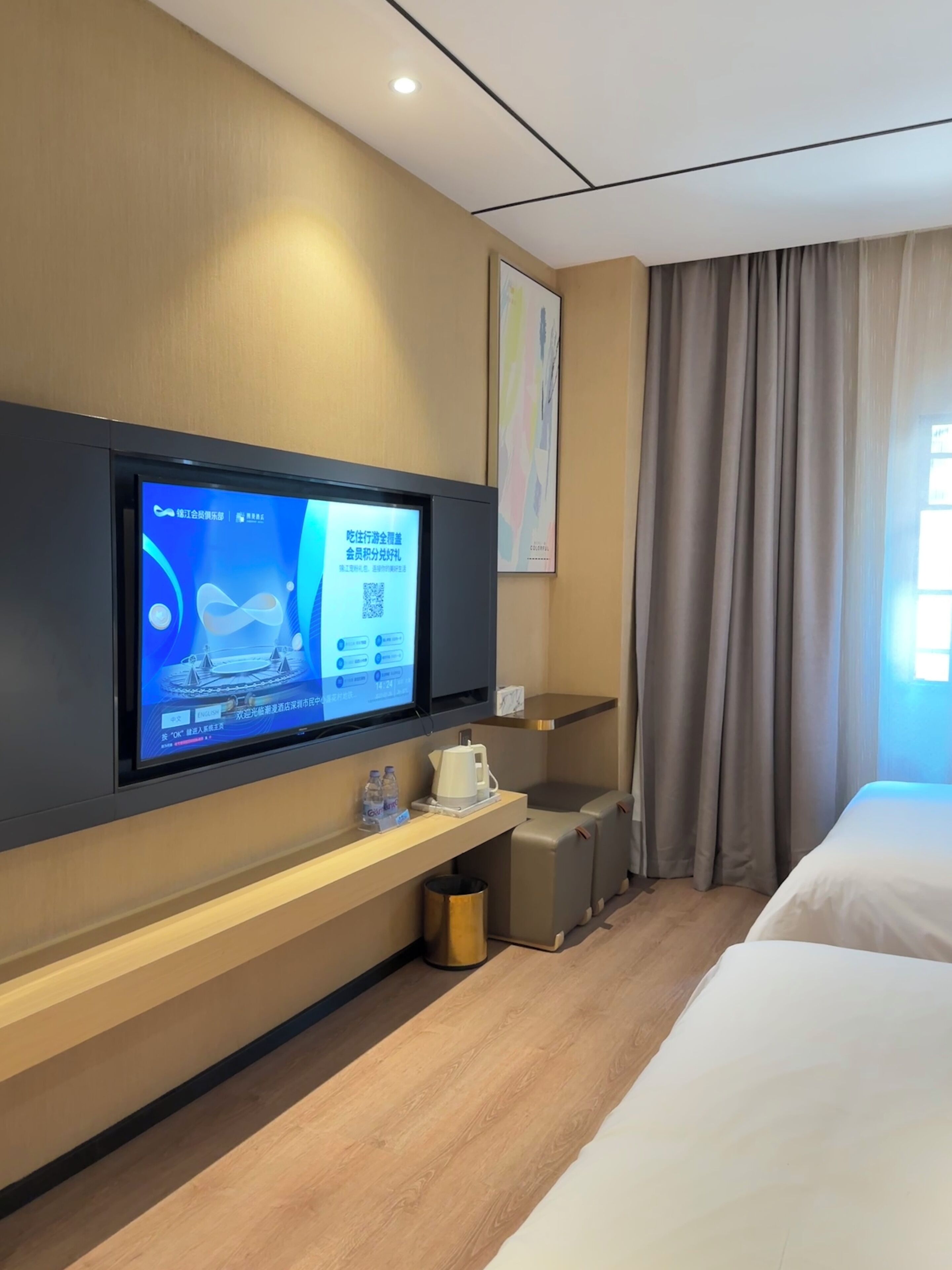 Ein modernes Hotelzimmer mit großem Flachbildfernseher an der Wand, einer kleinen Kaffeemaschine auf einem Regal, einem Mülleimer und einem Bett mit weißer Bettwäsche.