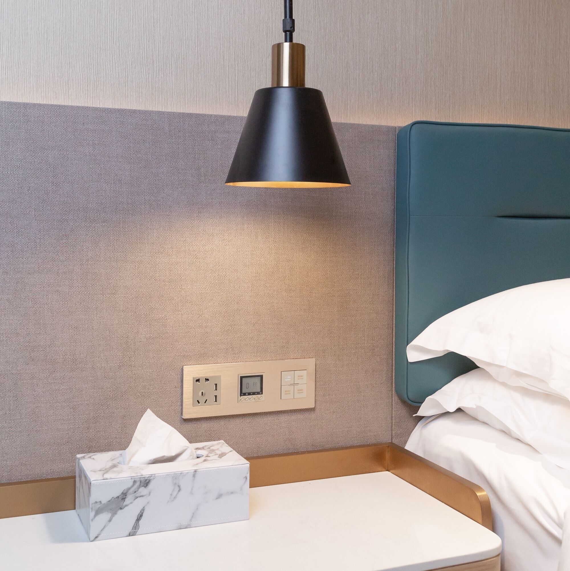 A hotel room with a bed, a bedside table, a marble tissue box, a wall socket, and a black pendant light.