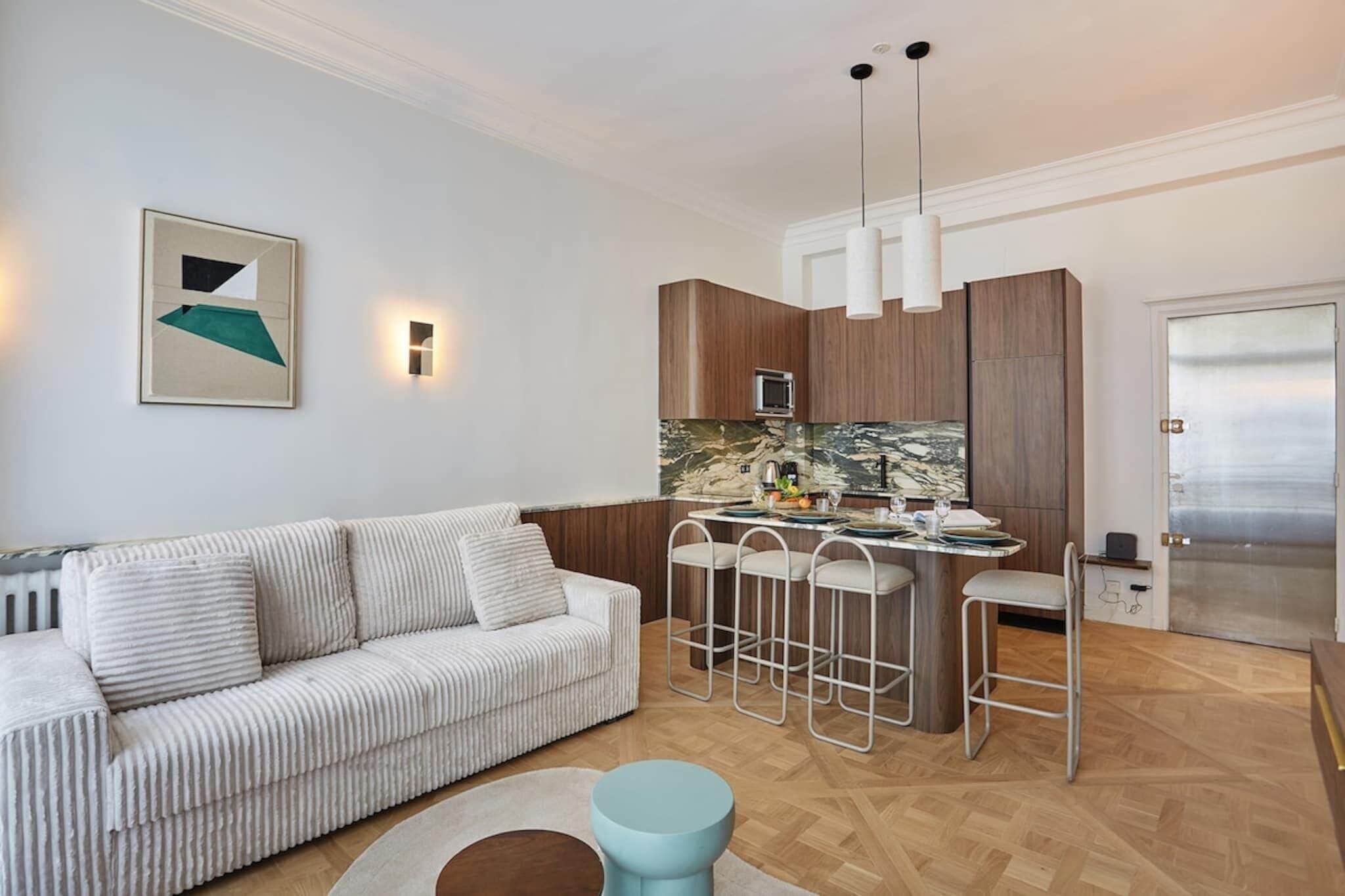 A modern living room with a striped sofa, a round coffee table, and a kitchen area with a bar stool and pendant lights.