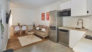 Een moderne keuken met een roestvrijstalen koelkast, een magnetron en een vaatwasser. Er is een eethoek met een kleine ronde tafel en twee witte stoelen. In de woonkamer staat een beige bank met kussens.