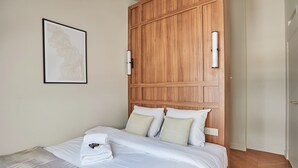 Una cama bien hecha con sábanas blancas y una toalla doblada, cabecera de madera y un cuadro en la pared.