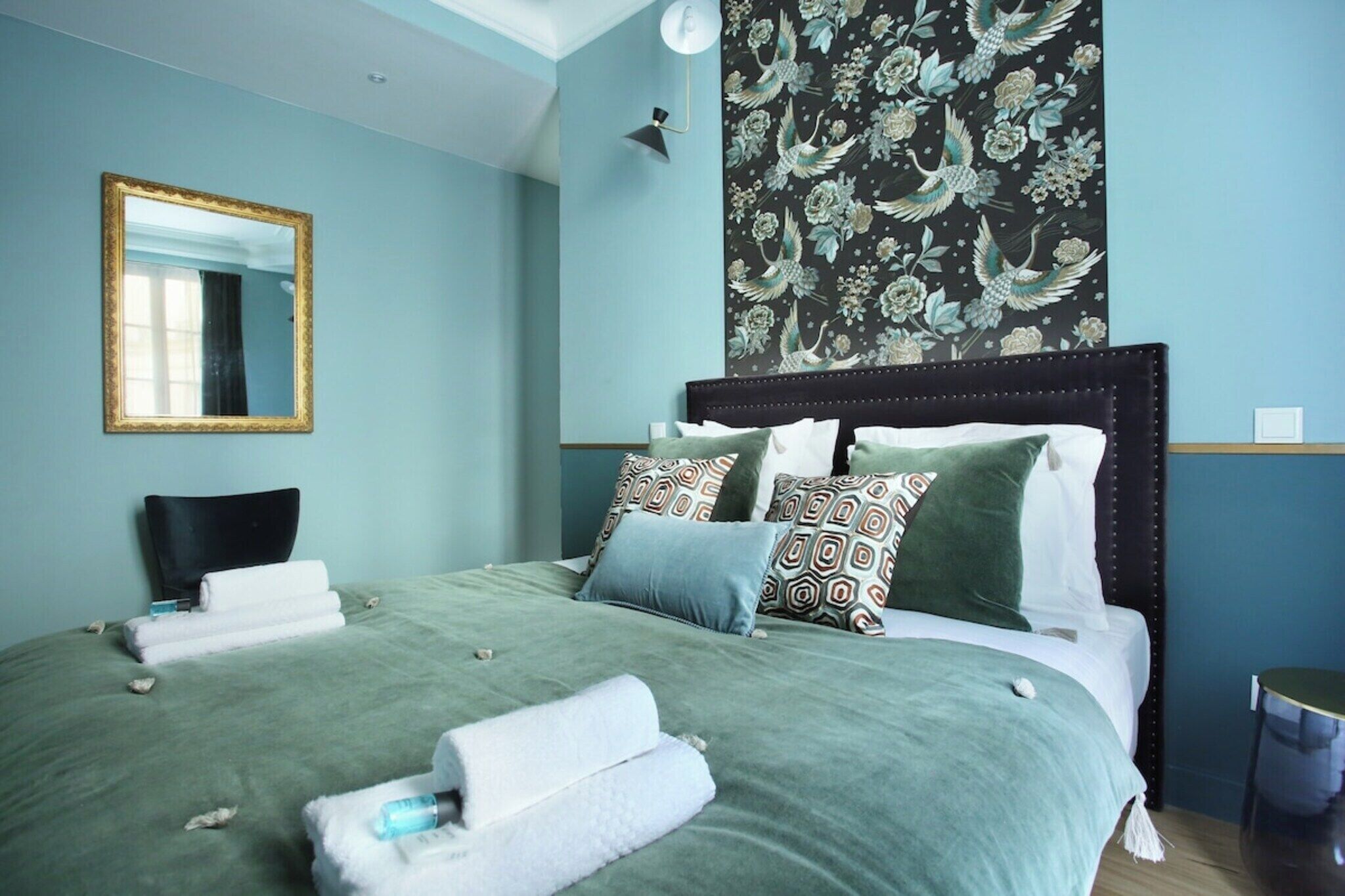 A neatly made bed with green bedding, a dark headboard, and decorative pillows. A large mirror with a gold frame is on the wall, and a floral tapestry hangs above the bed.