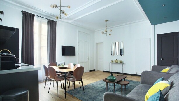 A modern living room with a grey sofa, a dining area with a round table and chairs, a kitchen area with a black countertop, and a large window with curtains. - Splendid apartment Champ de Mars - Eiffel Tower (Paris)