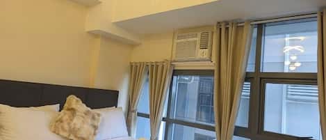 A hotel room with a bed, curtains, an air conditioning unit, and a window.