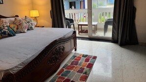 A hotel room with a large bed, a small table, a chair, a lamp, and a view of the outside balcony. - Apricot Inn (Morjim)
