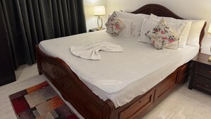 A neatly made bed with a white bedspread and floral pillows, a wooden headboard, a nightstand with a lamp, and a colorful rug. - Apricot Inn (Morjim)