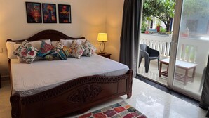 A neatly made bed with floral pillows, a wooden headboard, and a view of a balcony with outdoor furniture. - Apricot Inn (Morjim)