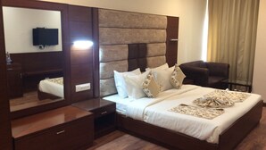 A modern hotel room with a large bed, a bedside table, a mirror, a TV, and a seating area with a chair and a glass table. - HOTEL THE SAFFRON PARK (Panchkula)