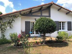 A white building with a tiled roof, a wooden porch, and a small tree in front.