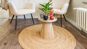 A room with two beige chairs, a round wooden table with a woven rug, a potted plant, a glass, and a bottle of pink grapefruit juice.