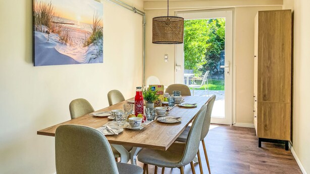 A dining area with a wooden table set for four, surrounded by chairs, a hanging light fixture, and a view of a garden through a glass door.