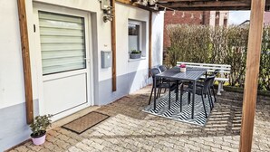 En overdækket terrasse med bord, stole, en dør og en lille plantepotte.