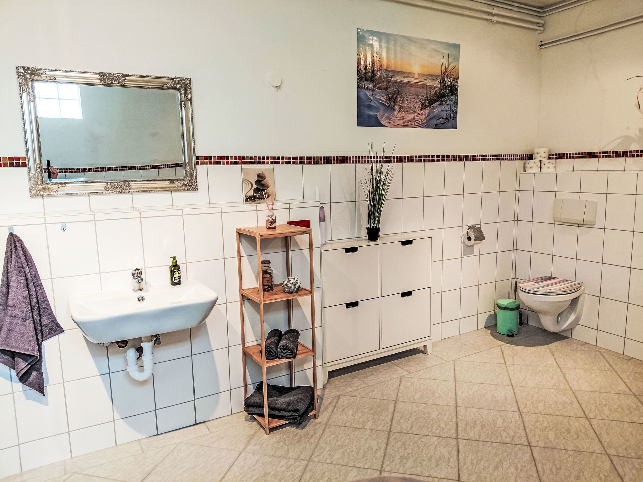 A bathroom with a white sink, a mirror, a wooden shelf with towels, a toilet, and a decorative picture on the wall.