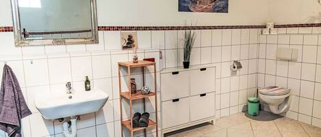 A bathroom with a white sink, a mirror, a wooden shelf with towels, a toilet, and a decorative picture on the wall.
