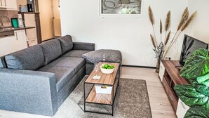 En moderne stue med en grå hjørnesofa, et træbord med en skål grønne æbler, et tv på et træmøbel og en stor grøn plante.