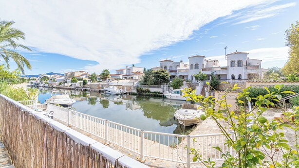 Un canal où naviguent des bateaux, une clôture blanche et des bâtiments aux toits recouverts de tuiles rouges.