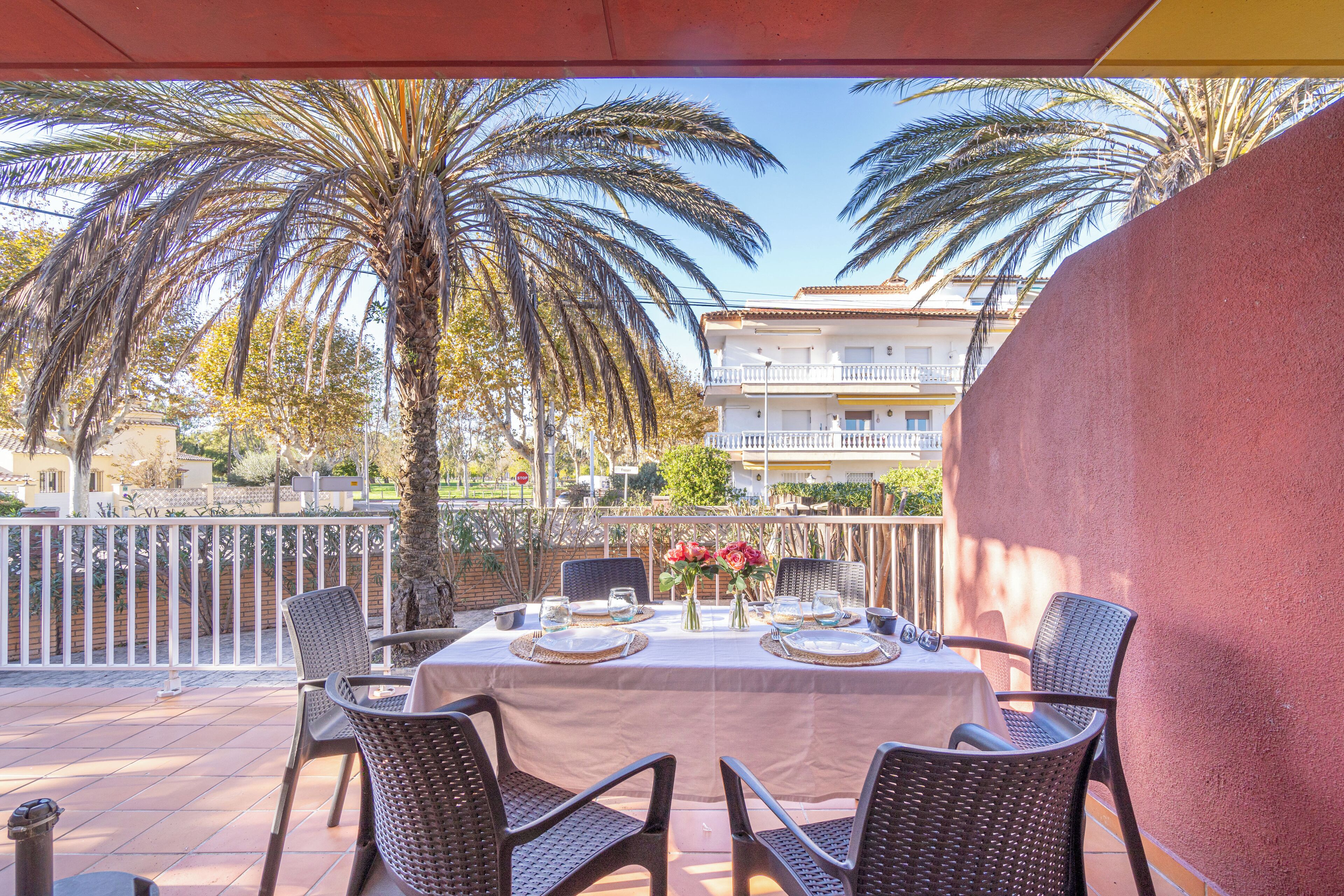 An outdoor patio with a table set for dining, surrounded by wicker chairs, palm trees, and a building in the background.