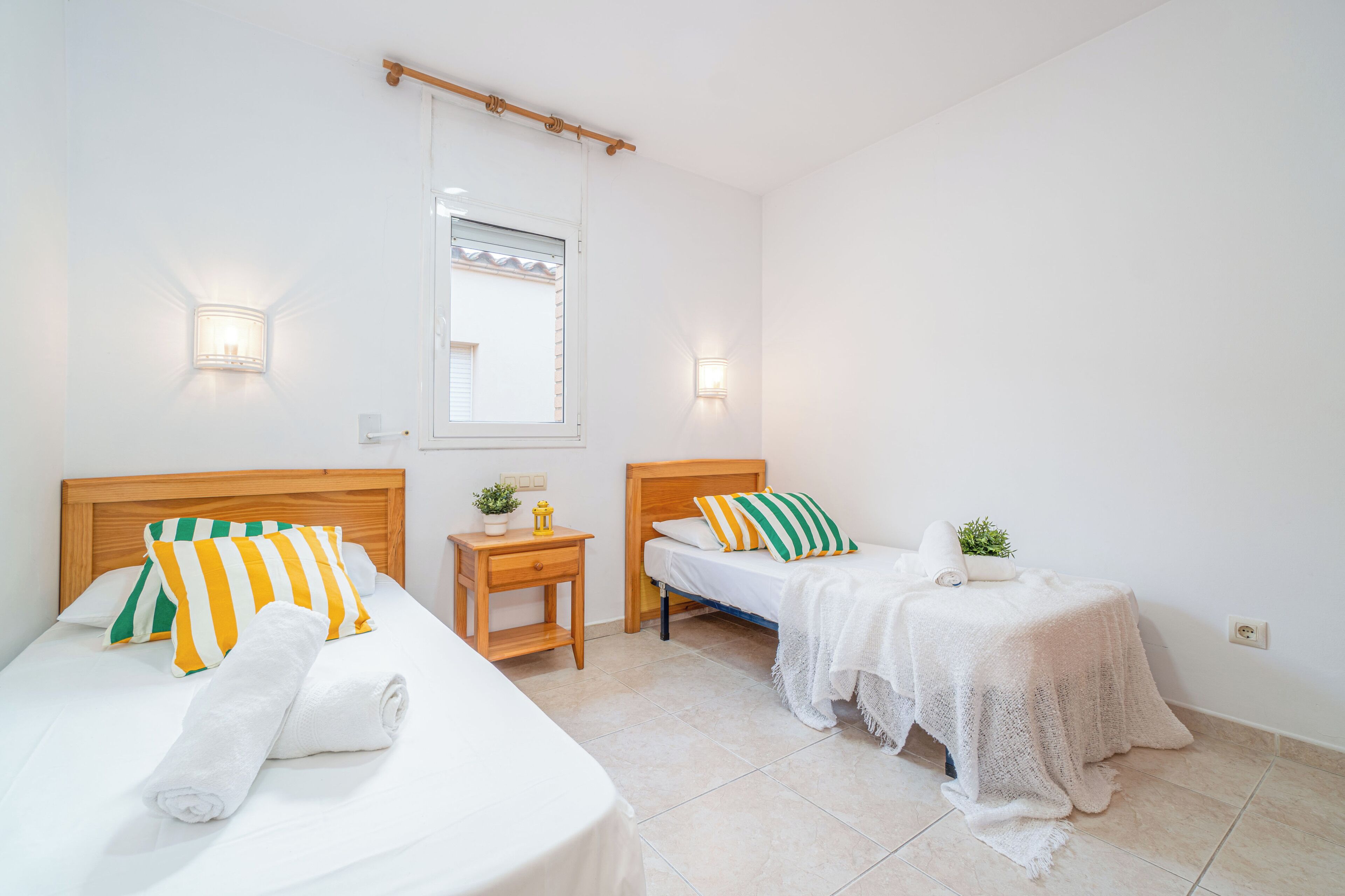 A bedroom with two beds, each with a wooden headboard, a small bedside table, and a wall-mounted light.