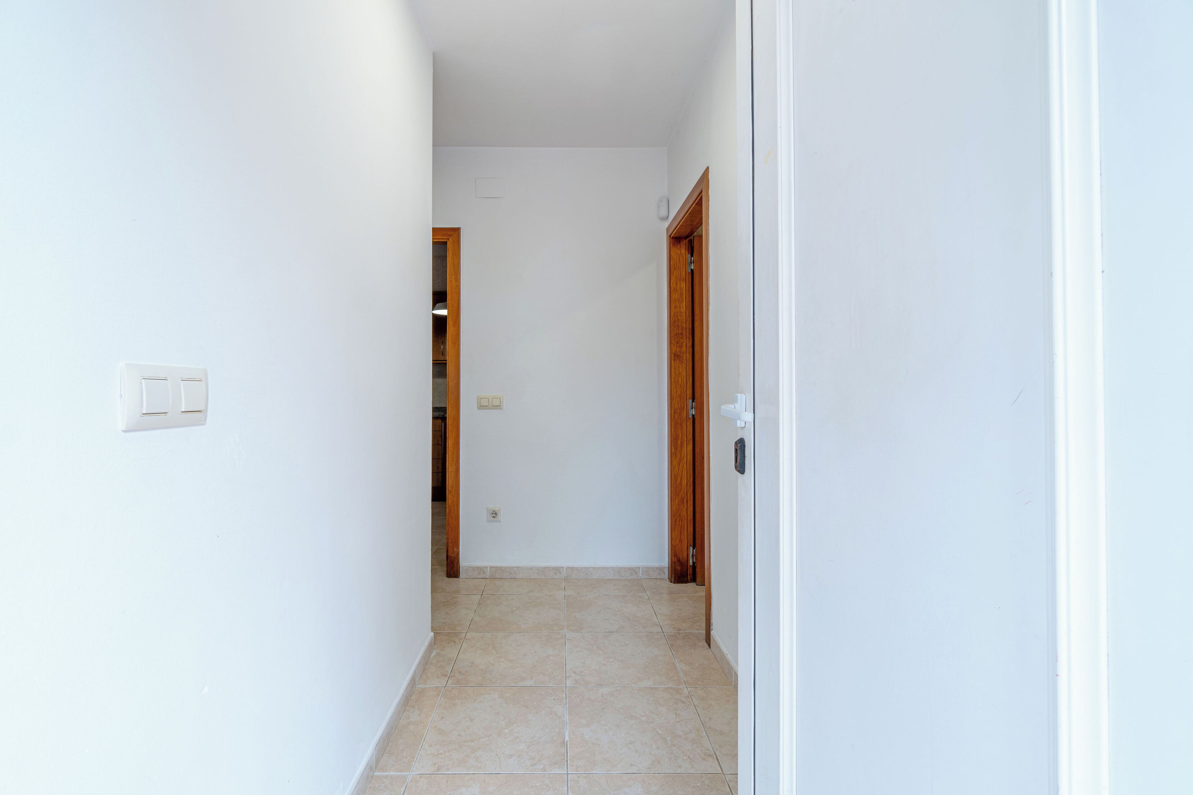A narrow hallway with white walls and a tiled floor. There are two wooden doors on the right, one of which is slightly ajar.
