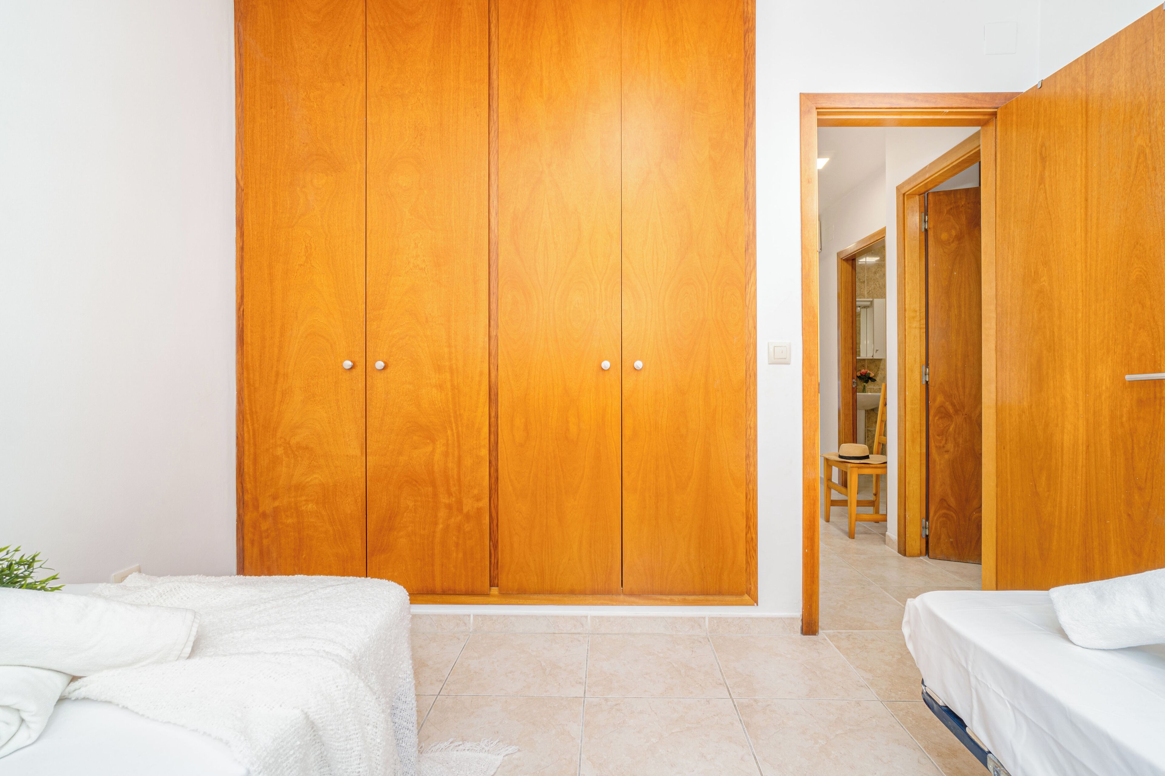 A bedroom with a large wooden wardrobe, a bed with white bedding, and a small table with a chair.