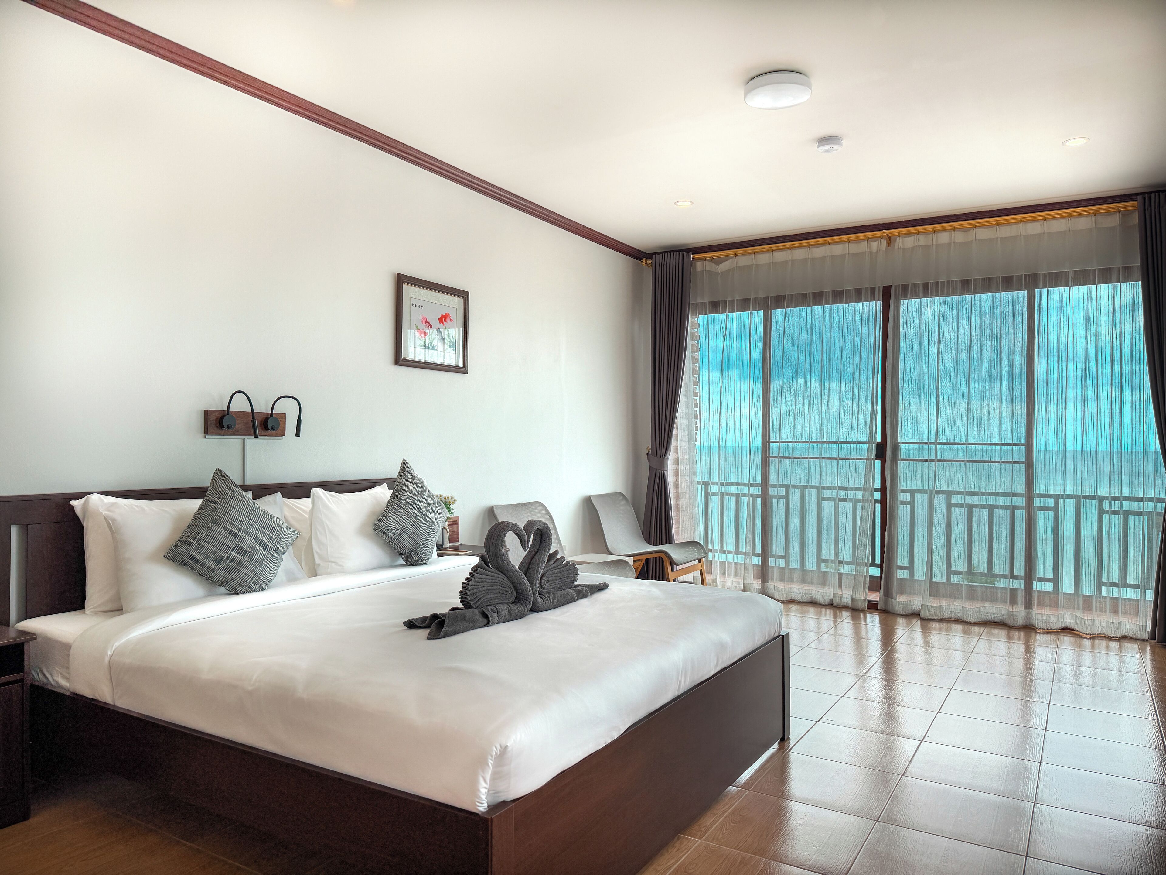 A hotel room with a large bed, a nightstand, a wall-mounted lamp, a framed picture, and a view of the outdoors through a balcony.