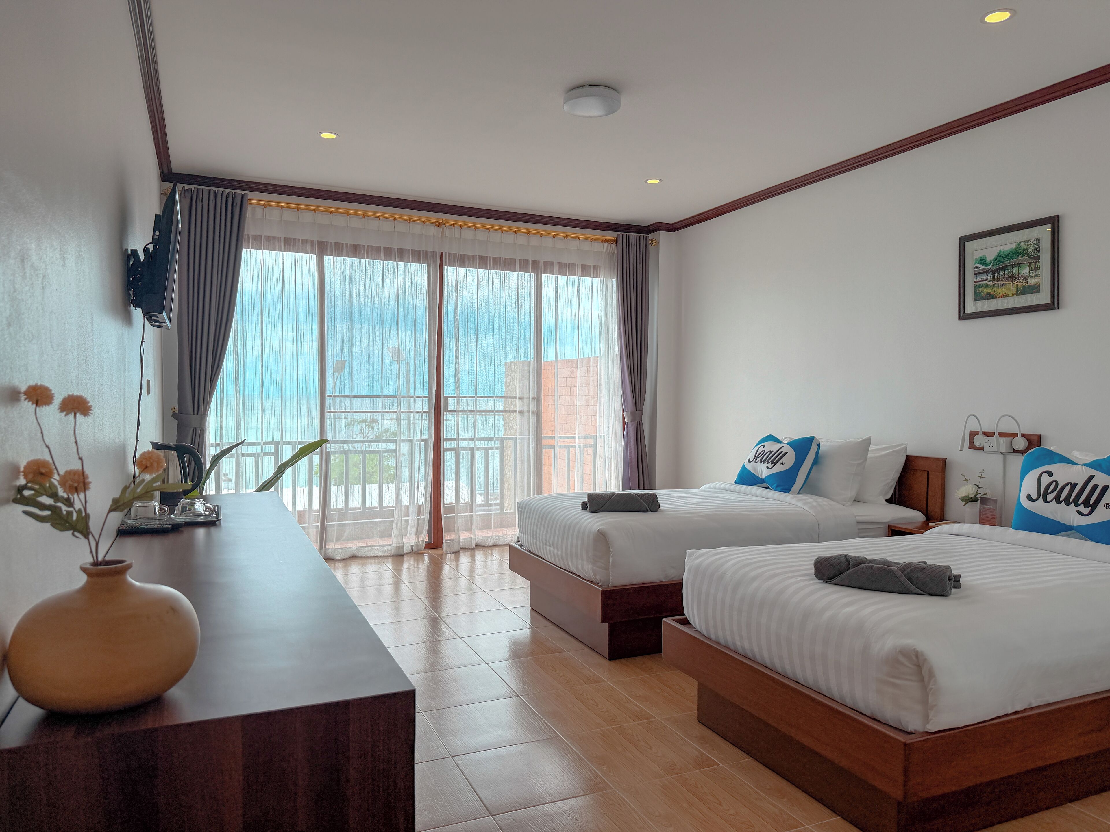 Habitación de hotel con dos camas, un escritorio con un florero, una ventana con cortinas y un cuadro en la pared.