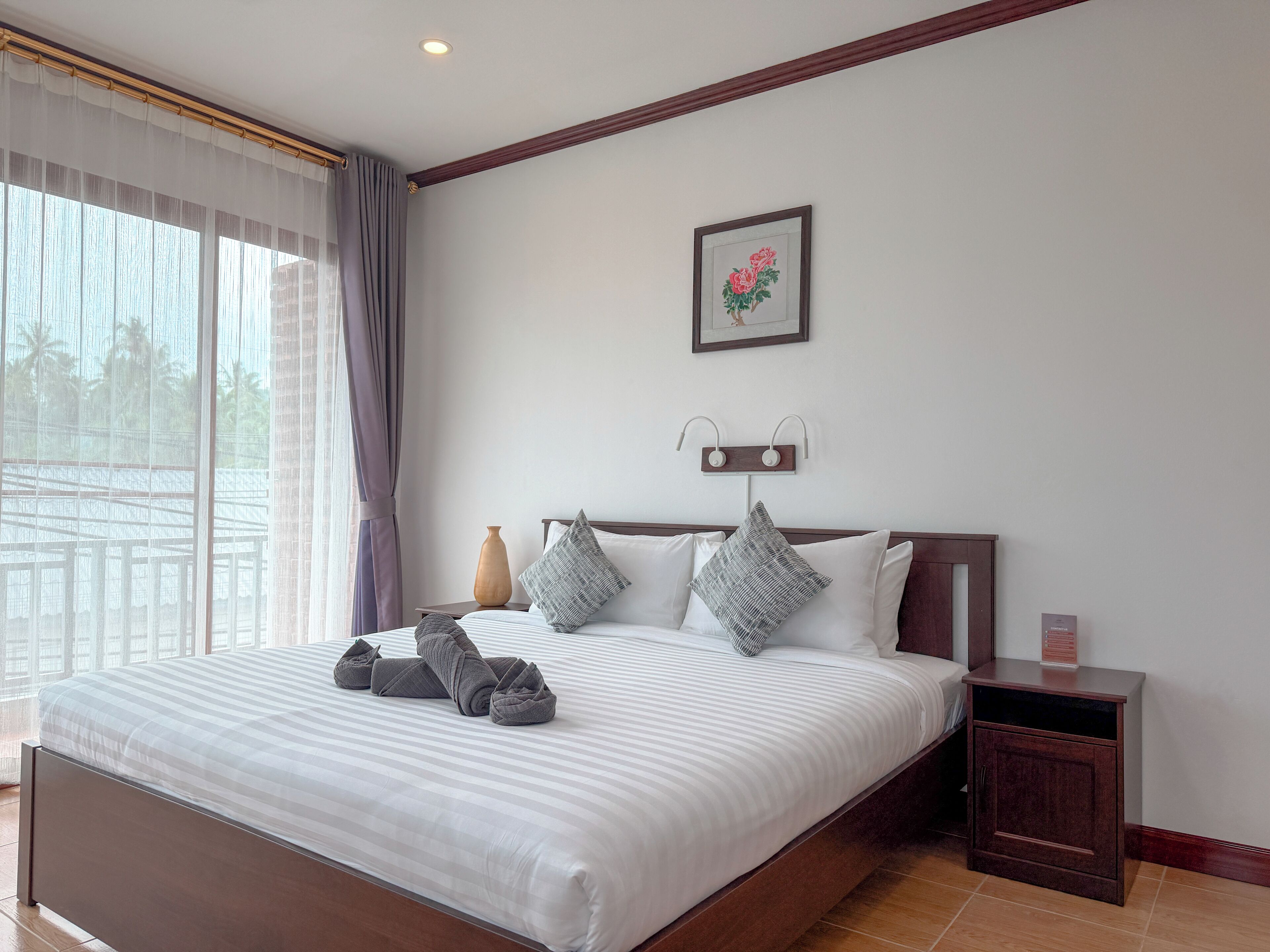 A neatly made bed with a striped white and gray duvet, a wooden nightstand with a vase, and a framed picture of flowers on the wall.