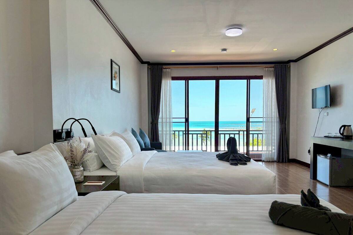 A hotel room with two beds, a desk with a TV, and a balcony with a view of the sea.