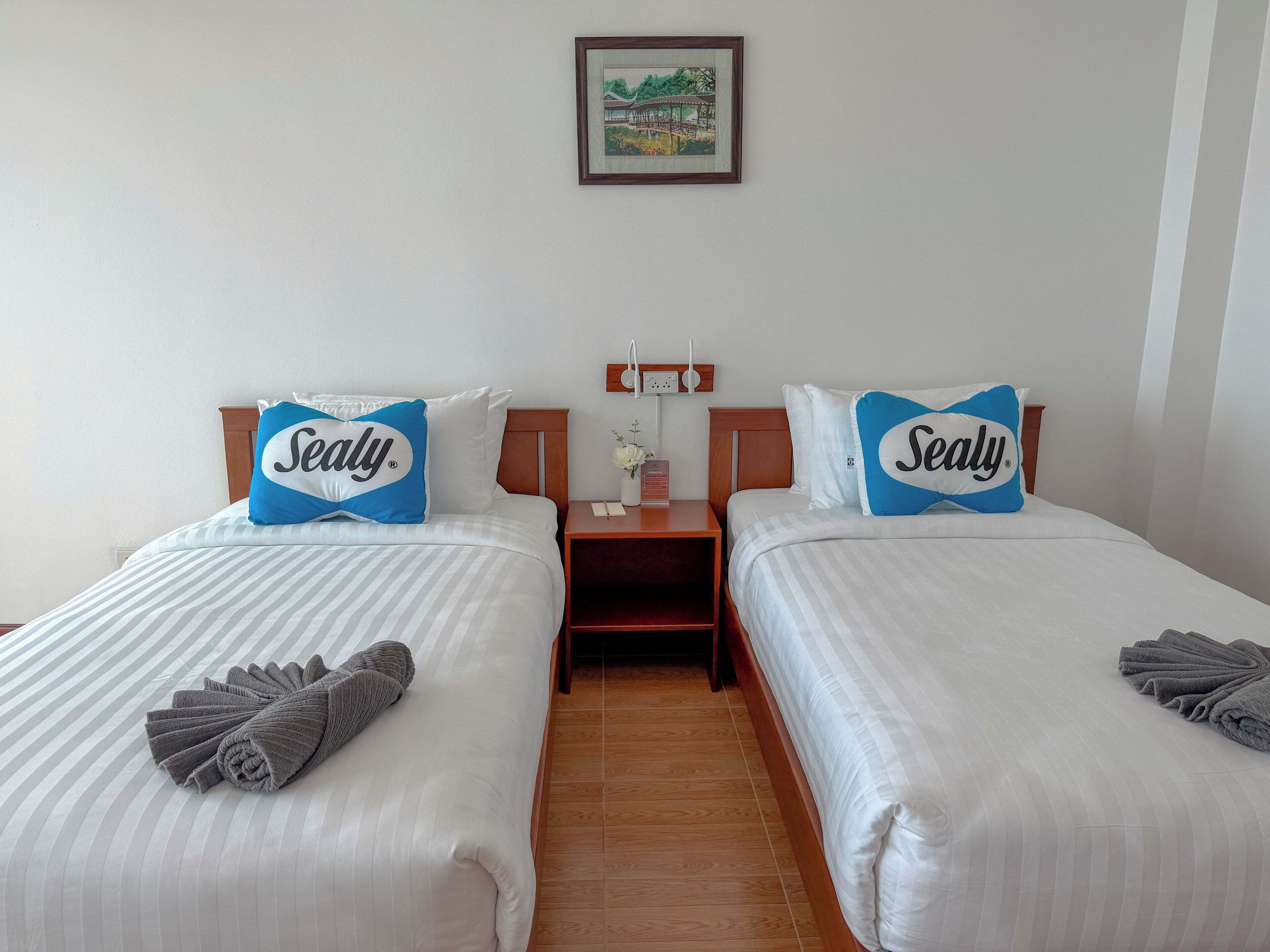 A hotel room with two beds, each with a Sealy branded pillow, a nightstand with a lamp, and a framed picture on the wall.