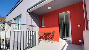 A balcony with a table and chairs, a glass door leading inside, and a red wall adjacent to a white building. - Charming studio near lake - Flower 42 (Zürich)