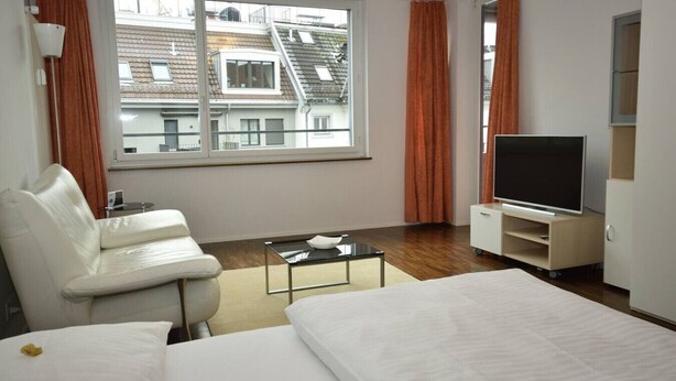 A hotel room with a bed, a white sofa, a TV on a stand, a small table, and a window with curtains. - Charming studio near lake - Flower 42 (Zürich)