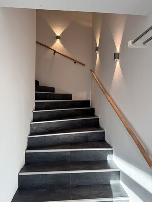 A modern staircase with dark stone steps and wooden handrails, illuminated by wall-mounted lights.