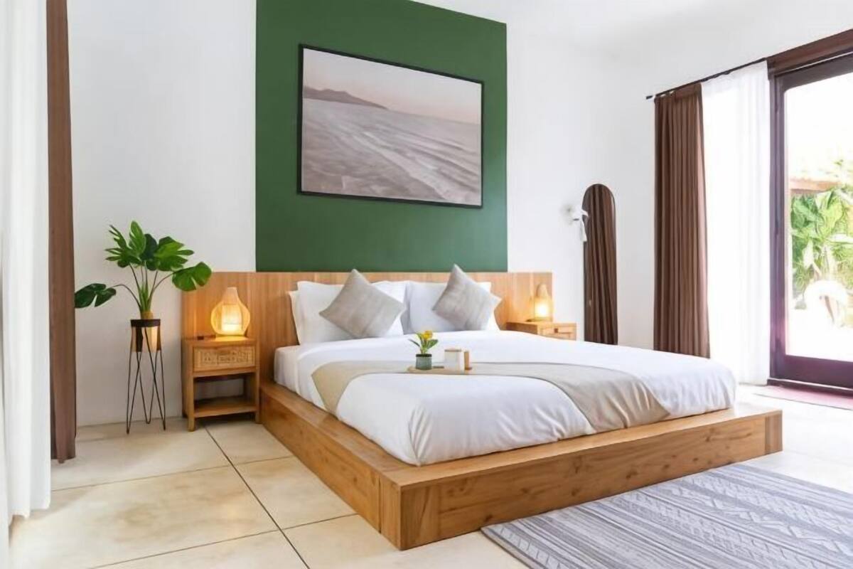 A bedroom with a wooden bed, a green accent wall, a framed picture, bedside lamps, and a potted plant.