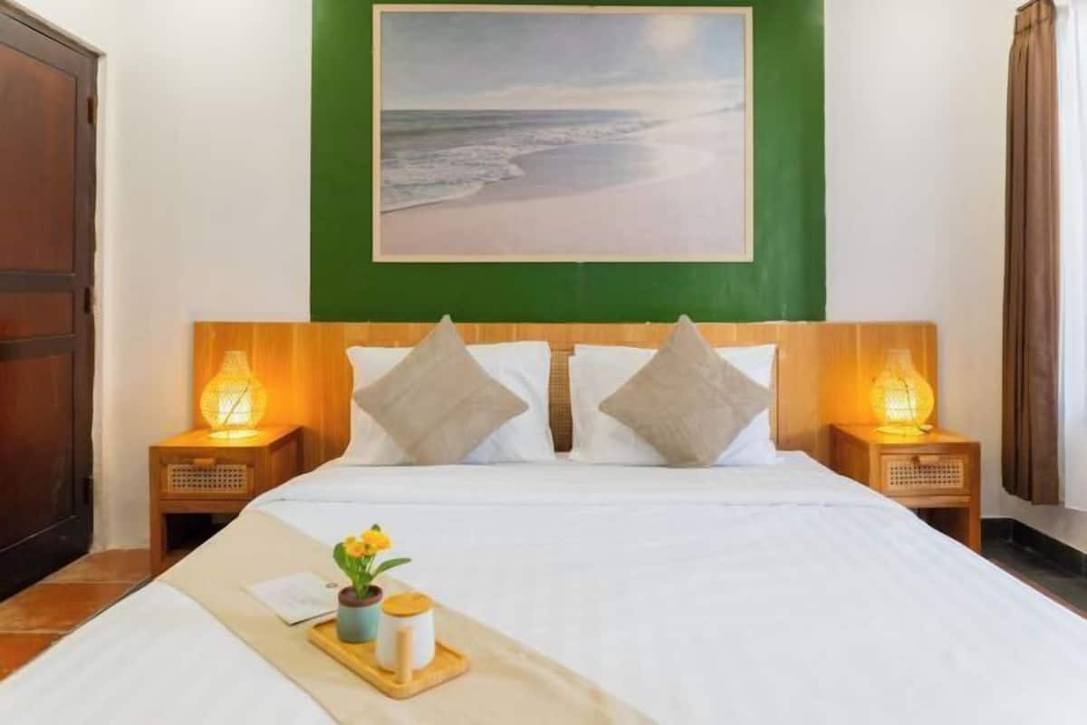 A hotel room with a bed, two bedside lamps, a painting of a beach, and a tray with flowers and a candle.