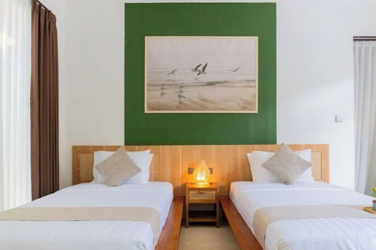 A hotel room with two beds, a nightstand with a lamp, a painting of birds on the wall, and a view of the beach.
