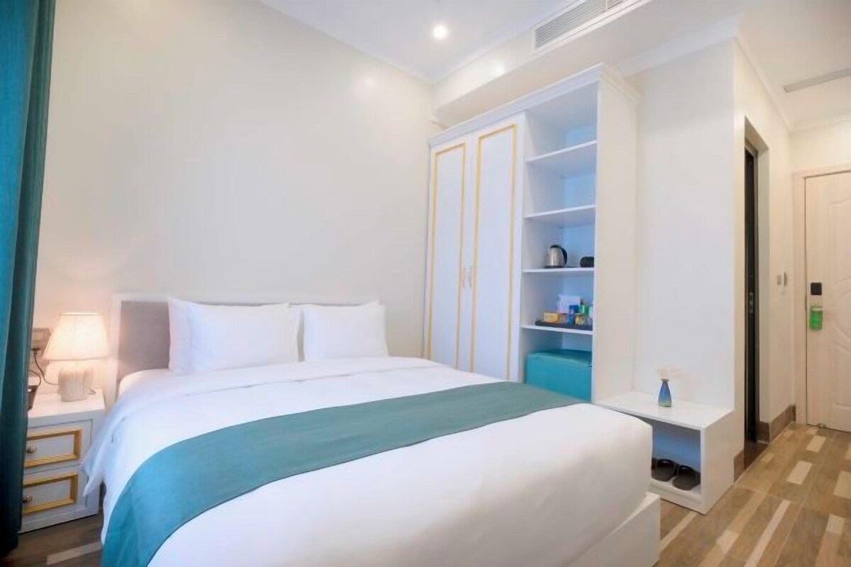 A hotel room with a bed, a nightstand, a wardrobe, and a small table with a lamp.