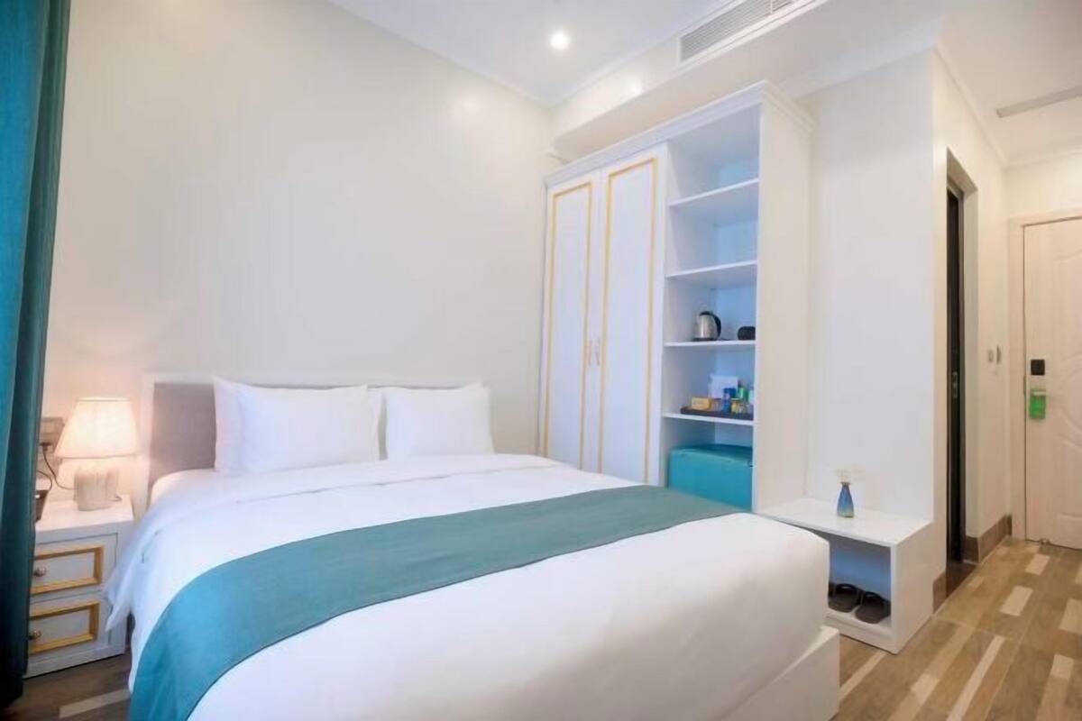 Habitación de hotel con cama, mesita de noche con lámpara, armario y una pequeña estantería con objetos.