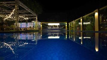 Una piscina nocturna con vista despejada al agua, una zona cubierta con guirnaldas de luces y un mural con cascada.