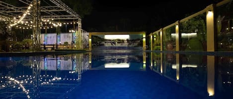 Une piscine nocturne offrant une vue dégagée sur l’eau, un espace abrité illuminé de guirlandes lumineuses et une fresque représentant une cascade.
