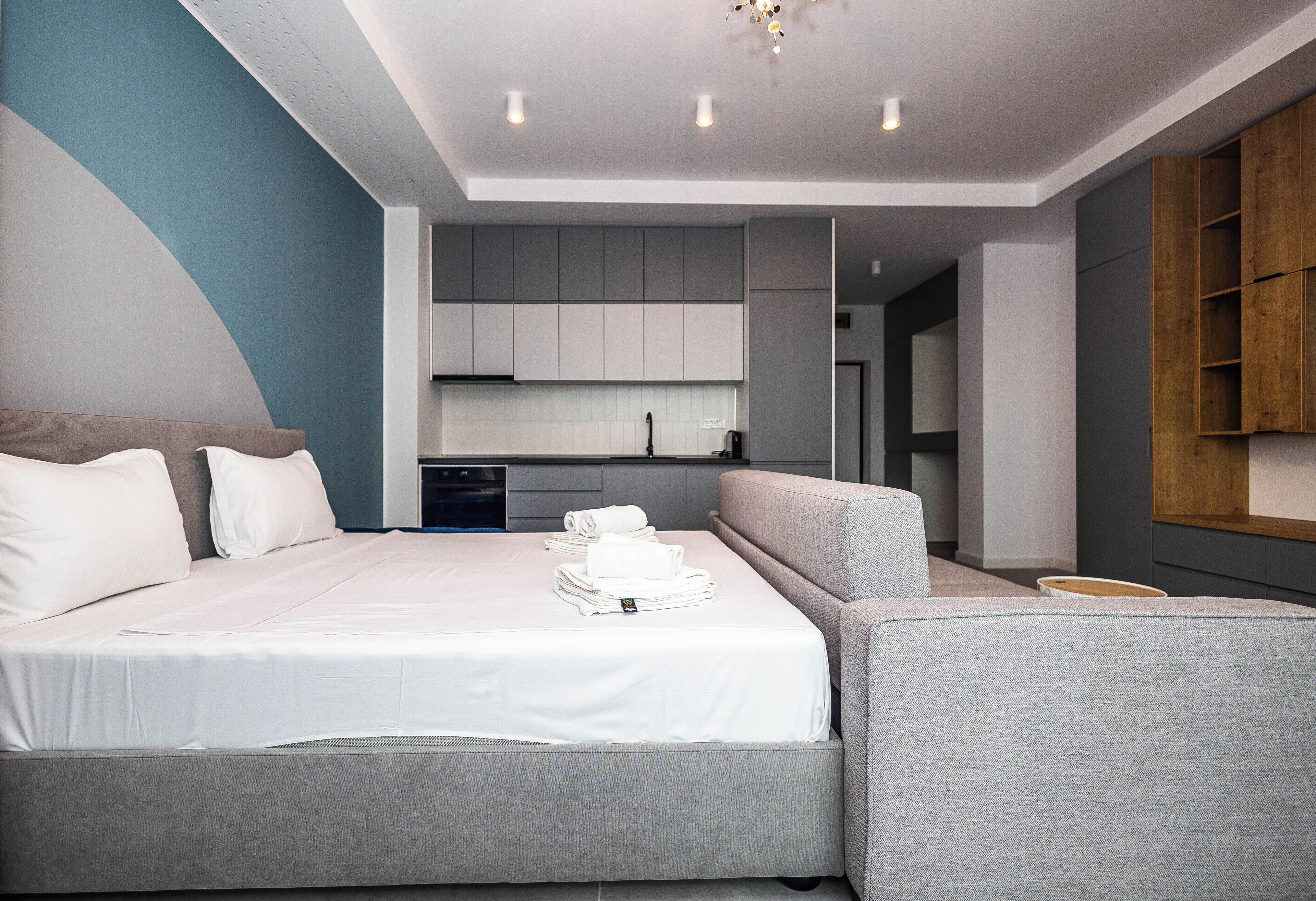 Quarto de hotel moderno com cama, sofá, uma mesinha e uma cozinha compacta ao fundo.