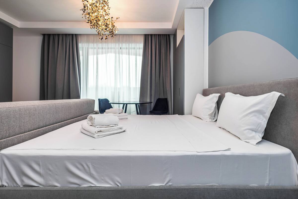 A modern hotel room with a large bed, grey upholstered headboard, white bedding, a small table with a chair, and a large window with sheer curtains.